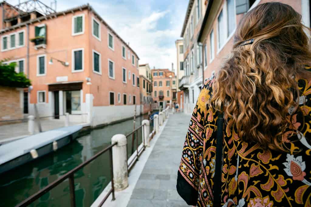 A local guide with long wavy hair walks along a canal in Venice, where orange buildings reflect on calm water under a partly cloudy sky.