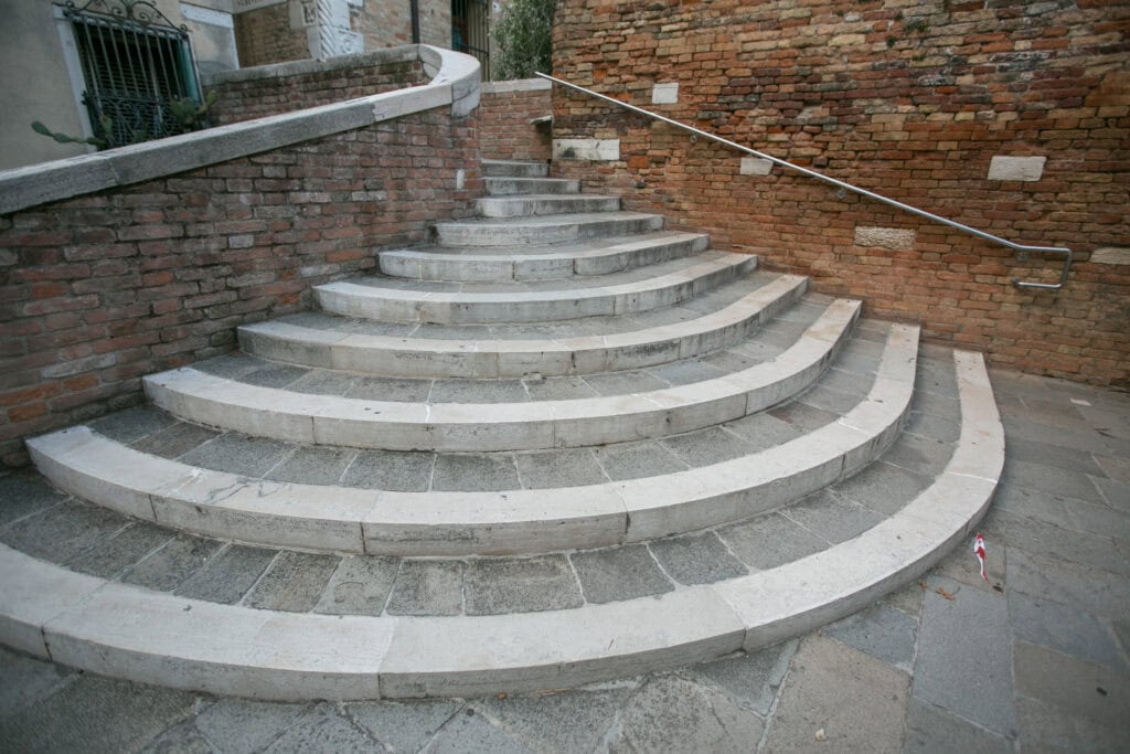 Wide stone steps curved in a semicircle, bordered by brick walls and a metal handrail, evoke the charm of local Venice pathways as they lead up to a higher walkway.