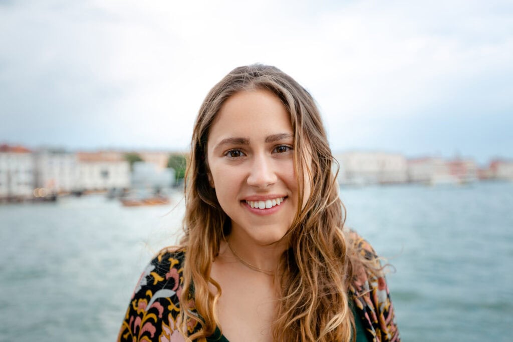 A young local woman with long wavy hair smiling, standing outdoors near a body of water with blurred Venice buildings in the background.