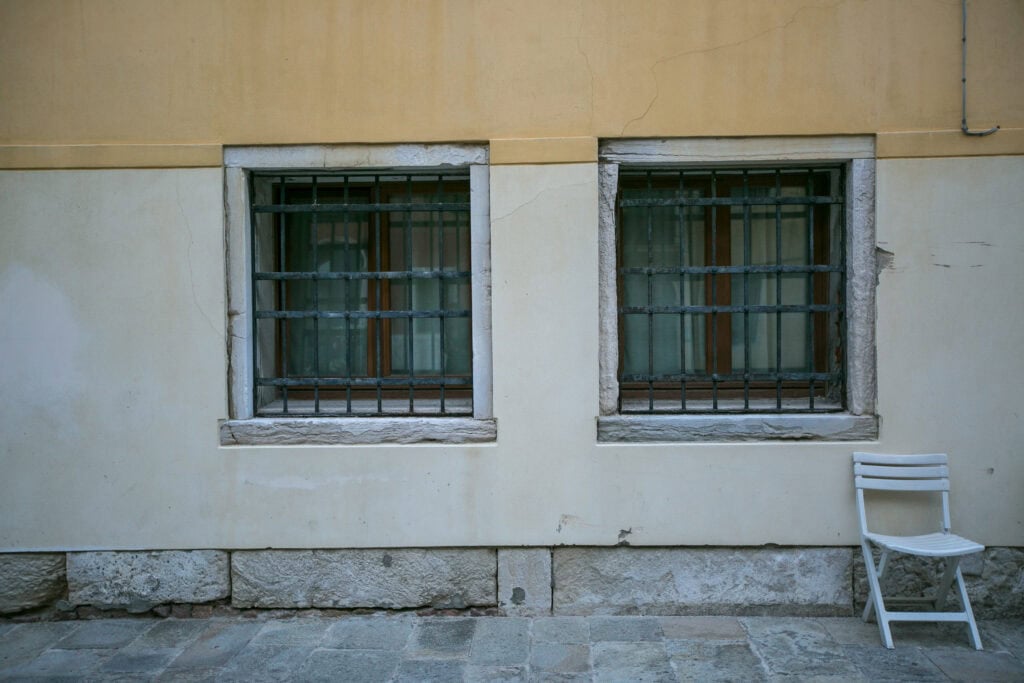 Two barred windows in a yellow and white wall above stone paving, with a single white folding chair to the right—capturing a quiet moment of Venice for those seeking an authentic local experience.
