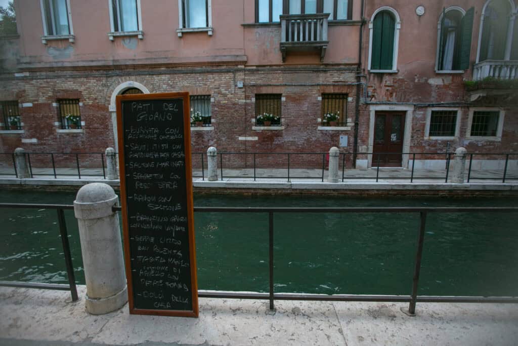 A chalkboard menu stands beside a canal in Venice, in front of an old brick building with arched windows and balconies, offering a charming local experience.