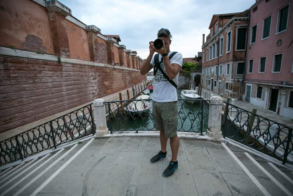 A person stands on a bridge over a canal in Venice, holding a camera and taking a photo; boats glide along the water below, capturing the essence of travel.