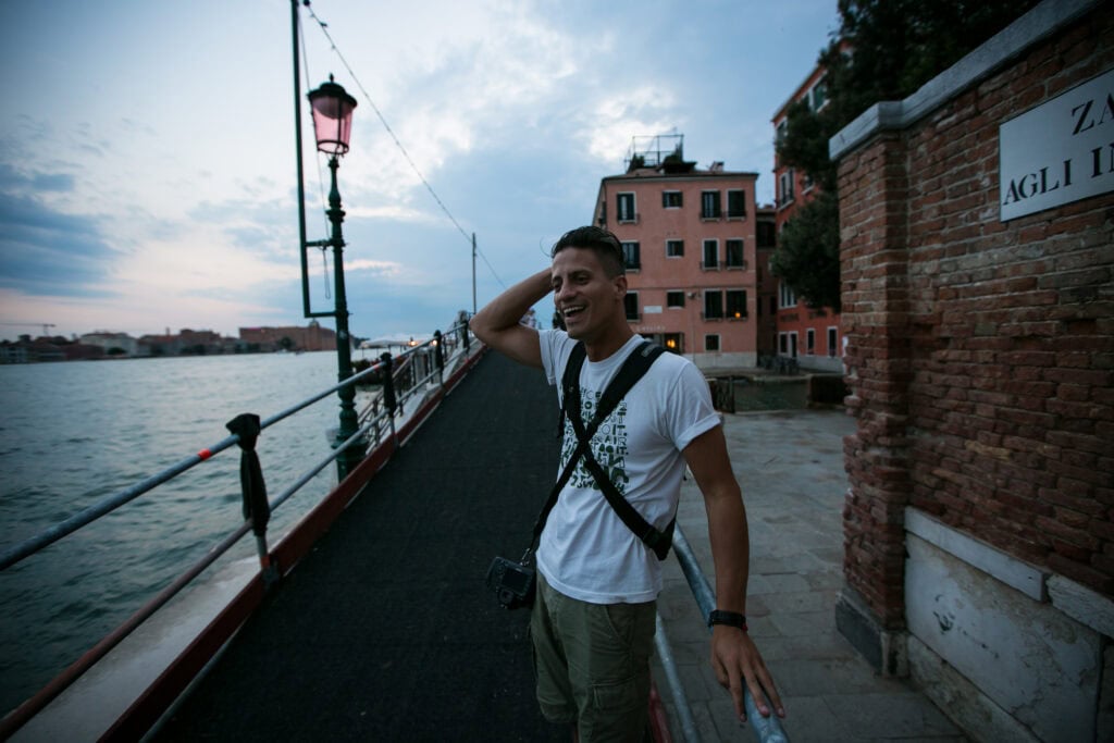 A man stands on a walkway by a canal in Venice, smiling with one hand on his head. Buildings and water are visible in the background under a cloudy sky, capturing the charm of travel in this iconic city.