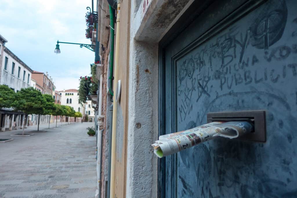 A rolled-up newspaper sticks out of a mail slot on a graffiti-marked door along an empty, narrow street lined with buildings and trees—an everyday scene waiting to be discovered by a curious traveler or local guide in Venice.