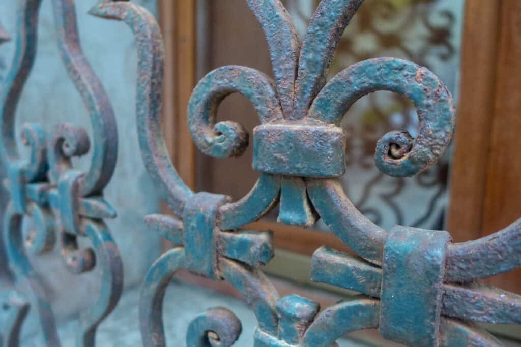 Close-up of an ornate, weathered metal fence with a blue-green patina and fleur-de-lis design, capturing the local charm of Venice, set in front of a wooden window.