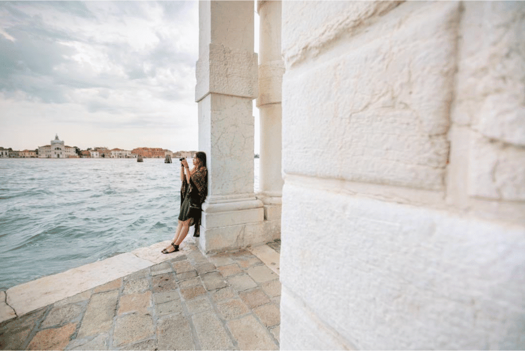 A person stands by a stone column near the water in Venice, taking a photo with a smartphone; local buildings are visible across the water under a cloudy sky.