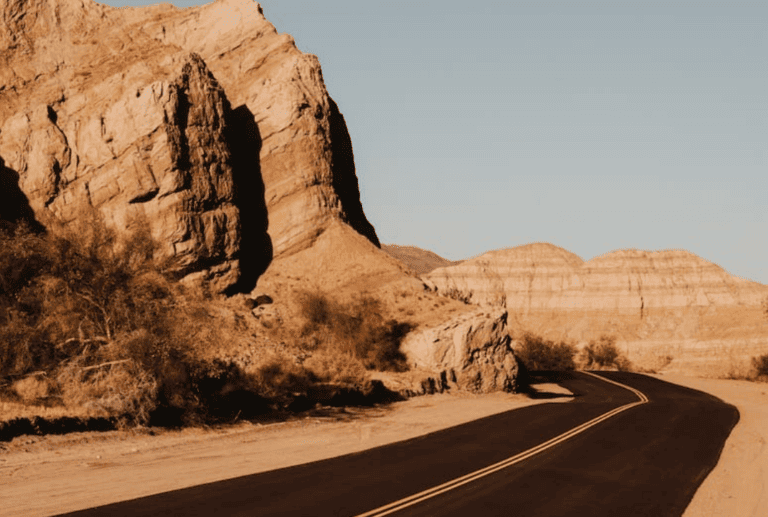 A paved road winds through a desert landscape with large rock formations and sparse vegetation under a clear sky, tracing the scenic route from San Diego to Palm Springs.