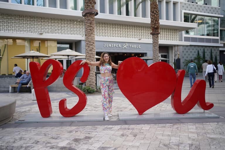 A woman posing in front of a heart-shaped sign at Palm Springs, perfect for an Instagram caption.
