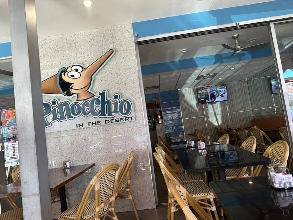 A café named "Pinocchio in the Desert," featuring a cartoon Pinocchio logo, with modern interior, empty rattan chairs, and glass-topped tables, known for offering the best breakfast