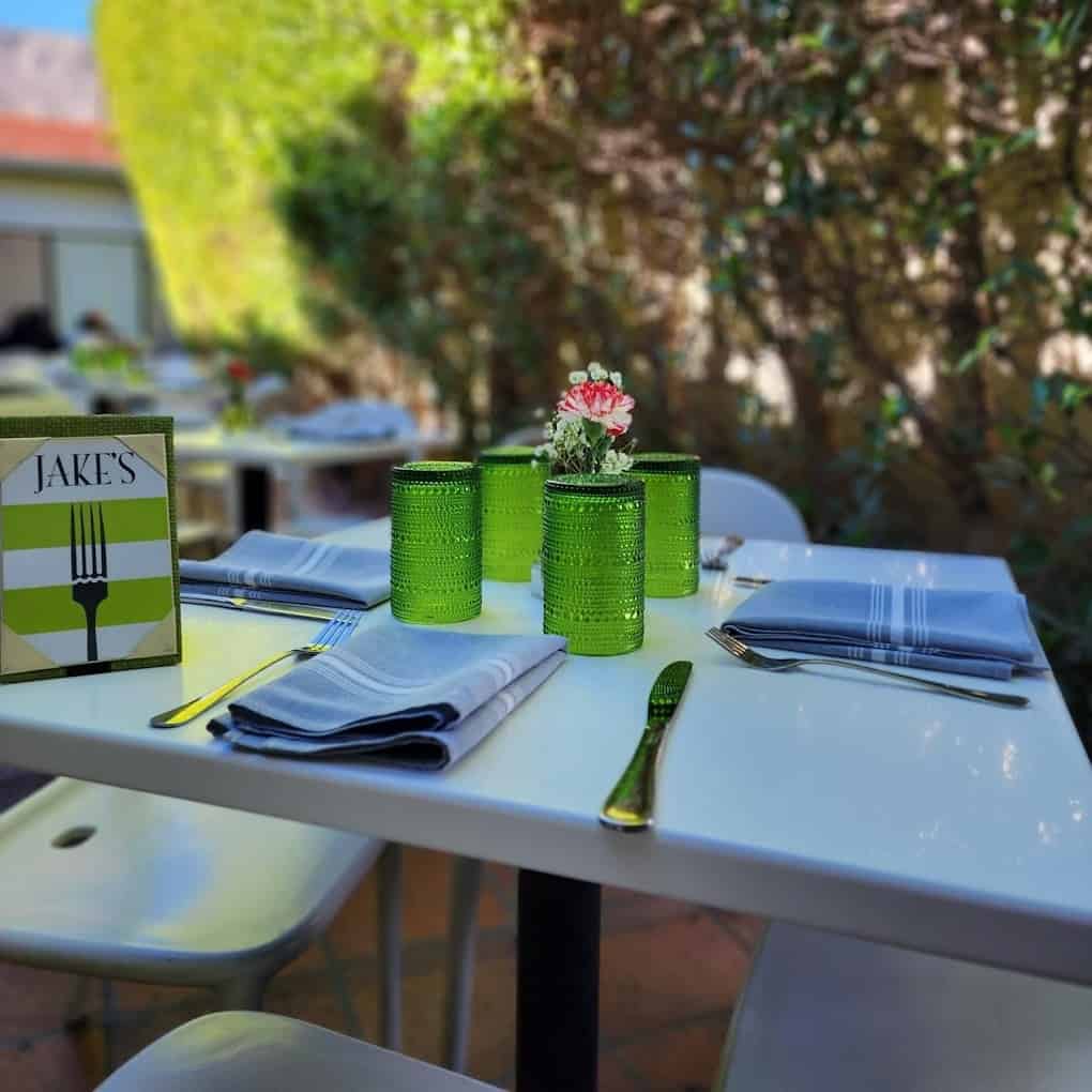Outdoor dining table set for two with green glass vases and a brunch menu labeled "Jake's" on a sunny patio with foliage in the background.