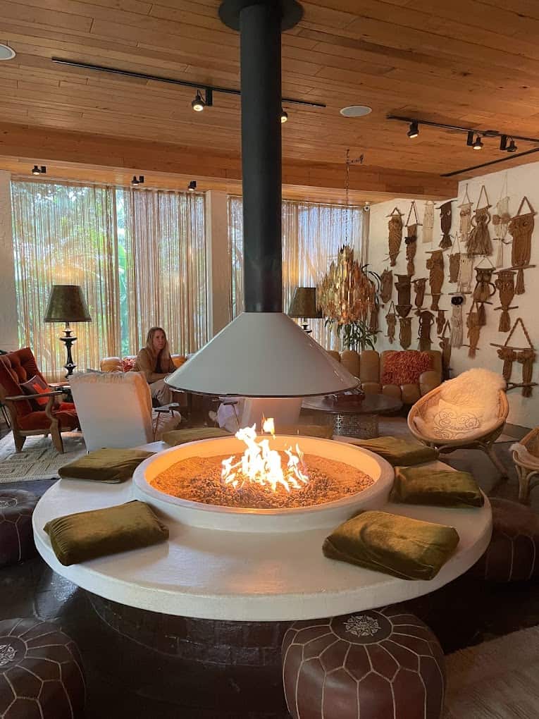 A woman sits in a cozy room with wood paneling, featuring a central circular fireplace, surrounded by cushions and eclectic wall decorations reminiscent of the best breakfast spots in Palm Springs.
