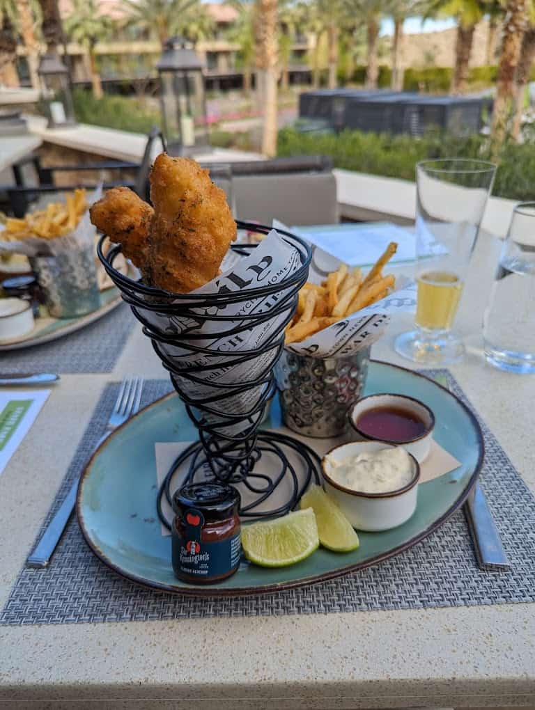 Fried fish and fries served in a spiral wire holder with lime wedges and dipping sauces on a blue plate, brunch in Palm Springs outdoor dining setting.