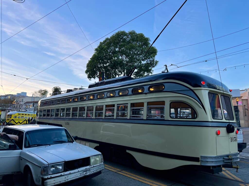 A vintage-style streetcar, reminiscent of the iconic cable cars in San Francisco, travels on tracks beside a parked white car and yellow vehicle. Overhead wires provide power, and a large tree is visible in the background under a blue sky.