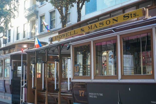 A Powell & Mason Sts. cable car is parked on a city street. The wooden car features an open seating area with benches and windows, typical of cable cars in San Francisco. Buildings and trees with lights are visible in the background.