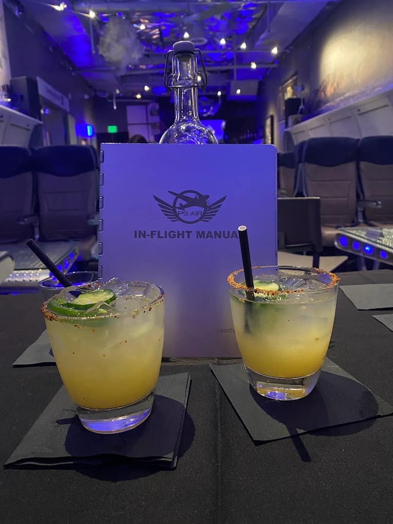 Two margaritas with lime garnish in front of an "in-flight manual" on a tray table inside an airplane cabin, evoking a brunch in Palm Springs ambiance.