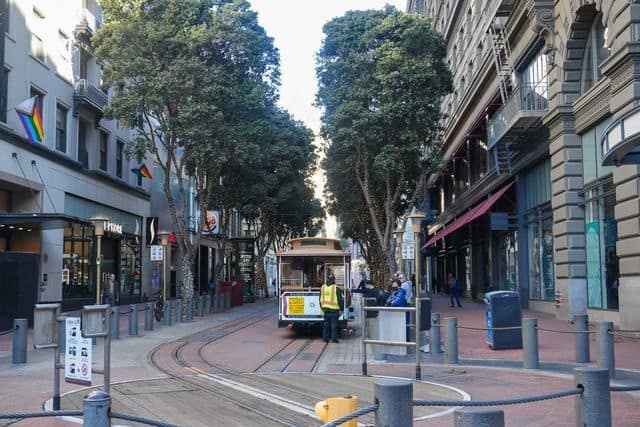 A narrow city street with shops on both sides, trees lining the road, and a classic San Francisco cable car in the center. Multicolored flags are displayed outside some stores. People are walking and sitting around.