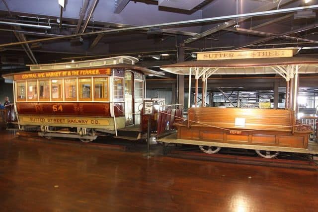 An old-fashioned streetcar labeled "Central Ave, Market St & The Ferries" and a connected open-air trolley labeled "Sutter St," reminiscent of the iconic Cable Cars in San Francisco, displayed in an indoor exhibit.