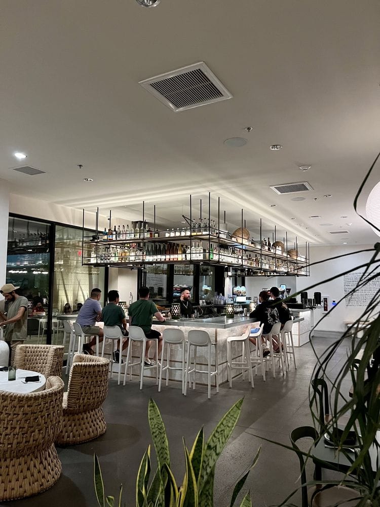 Modern bar and dining area with guests at tables, stools at the bar, ambient lighting, and greenery accents, ideal for brunch in Palm Springs.