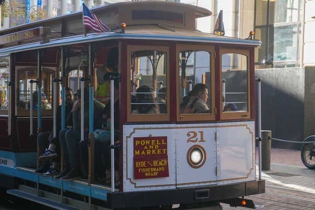 A Powell and Market streetcar, labeled "Hyde & Beach / Fisherman's Wharf," is pictured in motion with passengers inside. The streetcar, one of the iconic cable cars in San Francisco, has an American flag on top and the number 21 on its front.