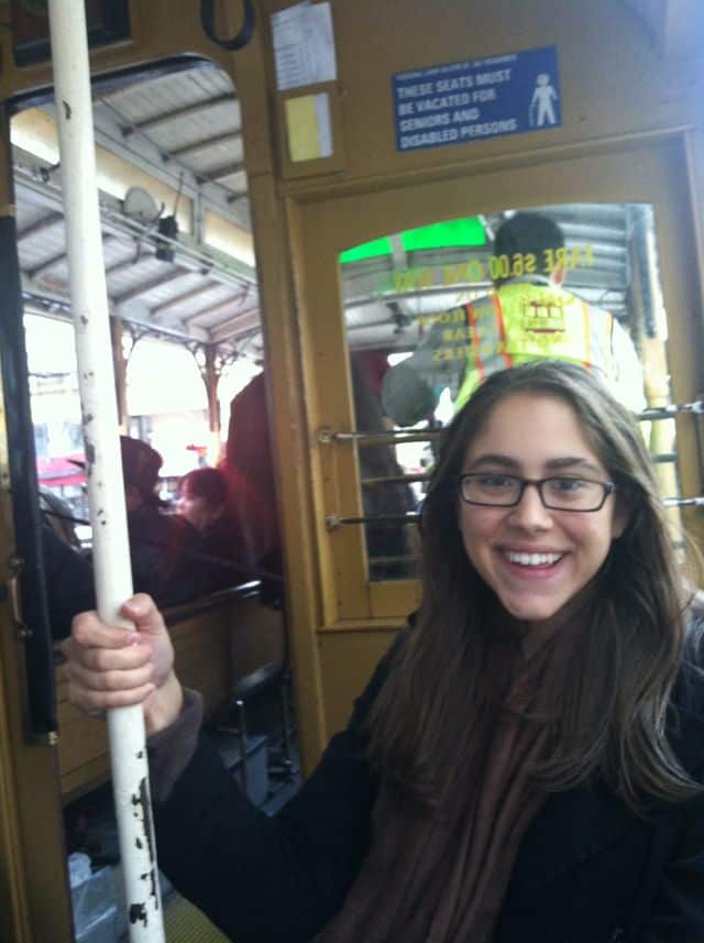 A smiling person with glasses and long hair holds onto a vertical pole inside a bus or cable car in San Francisco. A sign above their head indicates seats must be vacated for seniors and disabled persons.