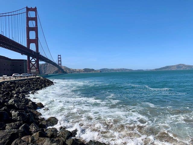 View of the Golden Gate Bridge from the rocky shoreline, with blue sky, ocean, and distant hills in the background, capturing a quintessential San Francisco scene where cable cars often weave through charming streets nearby.