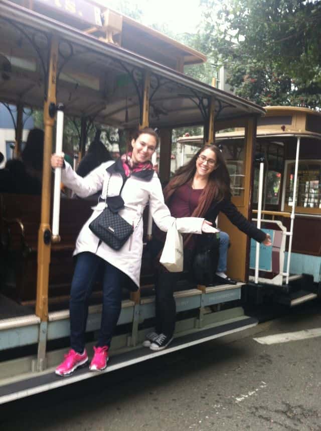Two women smiling and posing on the steps of a moving streetcar in San Francisco, one dressed in a white coat and pink sneakers, the other in a burgundy top and black sneakers. Trees are visible in the background, capturing the charm of cable cars in San Francisco.