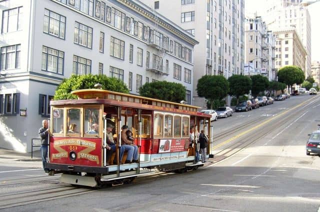 A red and cream cable car travels up a steep street in San Francisco, with people riding inside and others standing on the sides. Buildings and parked cars line the street.