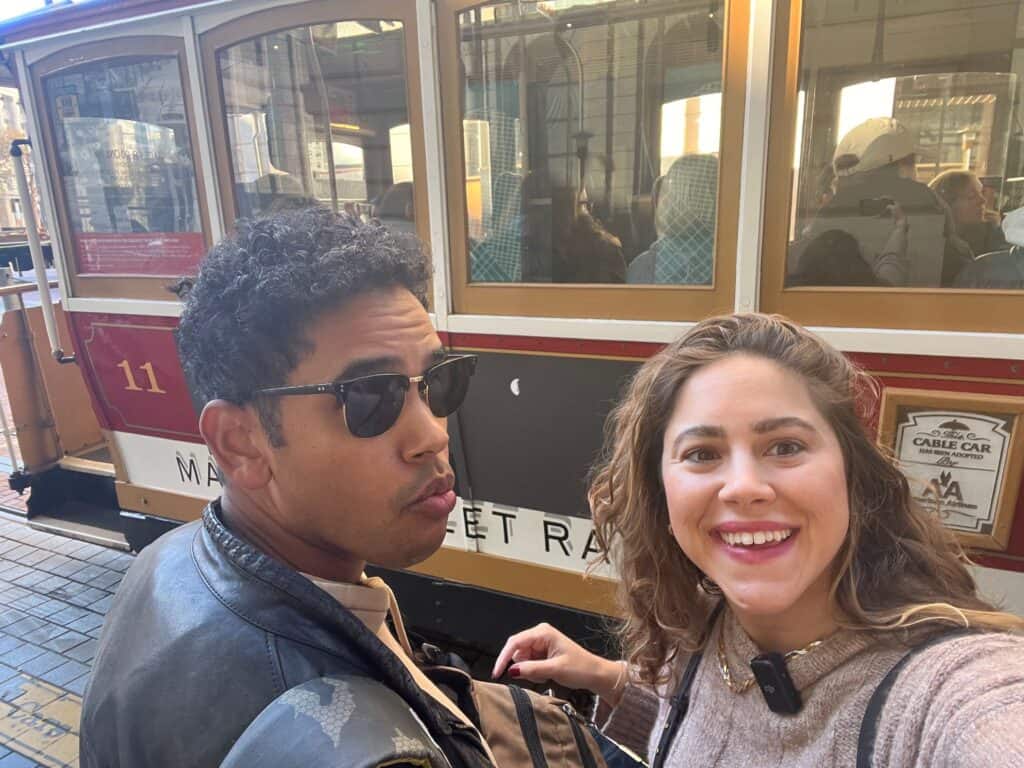 Two people, one wearing sunglasses and a leather jacket and the other smiling, taking a selfie in front of a red and gold cable car marked with the number 11, showcasing the iconic Cable Cars in San Francisco.
