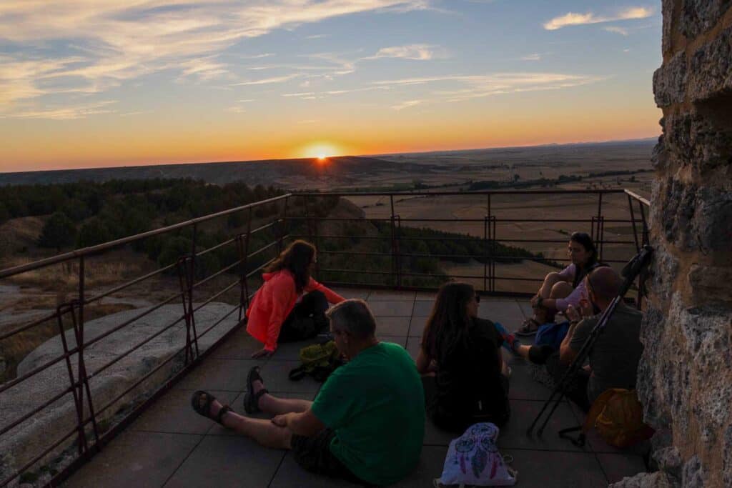 Five people sitting on a platform, looking at a sunset over a vast landscape with fields and trees. There is a stone wall on the right and a railing along the edge of the platform. They share Camino de Santiago tips while soaking in the serene beauty.