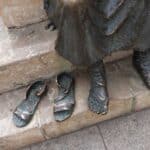 A surprising bronze statue of a standing figure with its sandals placed neatly on the steps beside its feet. The statue, situated on a stone staircase, offers an unexpected homage to the Camino de Santiago.