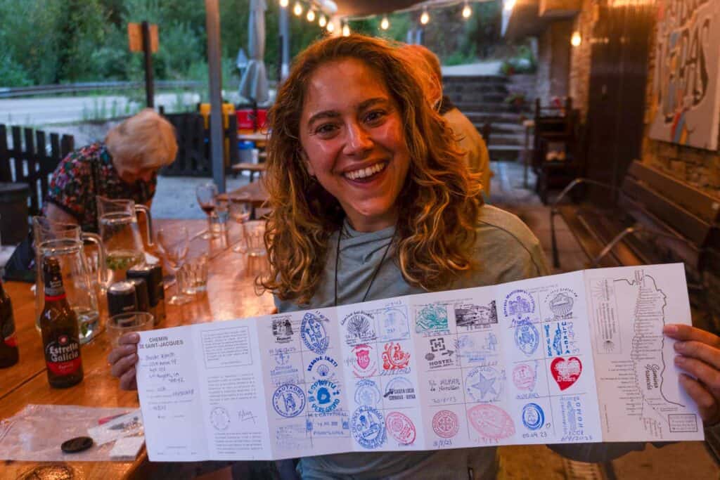 A person sits at an outdoor table holding an unfolded document filled with various stamps and smiling, likely from their journey. Empty glasses and a bottle are on the table, while several people are visible in the background, perhaps sharing Camino de Santiago tips.