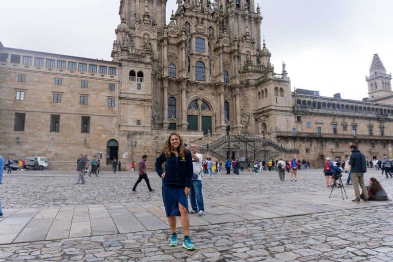 A woman in a blue outfit stands on a cobblestone square in front of a large historic cathedral, reminiscent of the scenes on the Camino de Santiago—an iconic pilgrimage route. Other people walk and sit in the background, adding to the lively atmosphere.