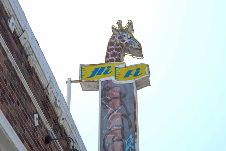 A vintage neon sign featuring a giraffe head with sunglasses and the words "Hi Fi" on a yellow banner attached to a building.