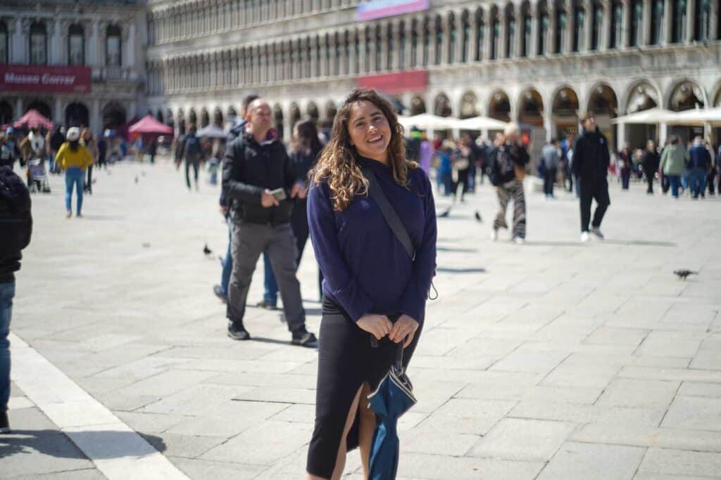 A woman in a blue top and black skirt stands smiling in a busy square with a historic building and several people in the background, possibly reflecting on her recent GigSky review.