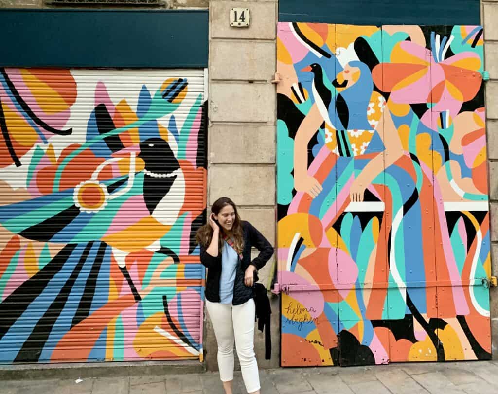 A person stands smiling in front of a brightly colored mural featuring abstract birds and human figures on two large doors, capturing the vibrant spirit of Barcelona.
