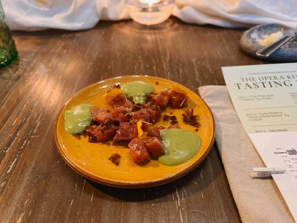 A small portion of tuna tartare with green sauce and garnish served on an orange plate. A tasting menu and napkin are partially visible on the wooden table nearby.