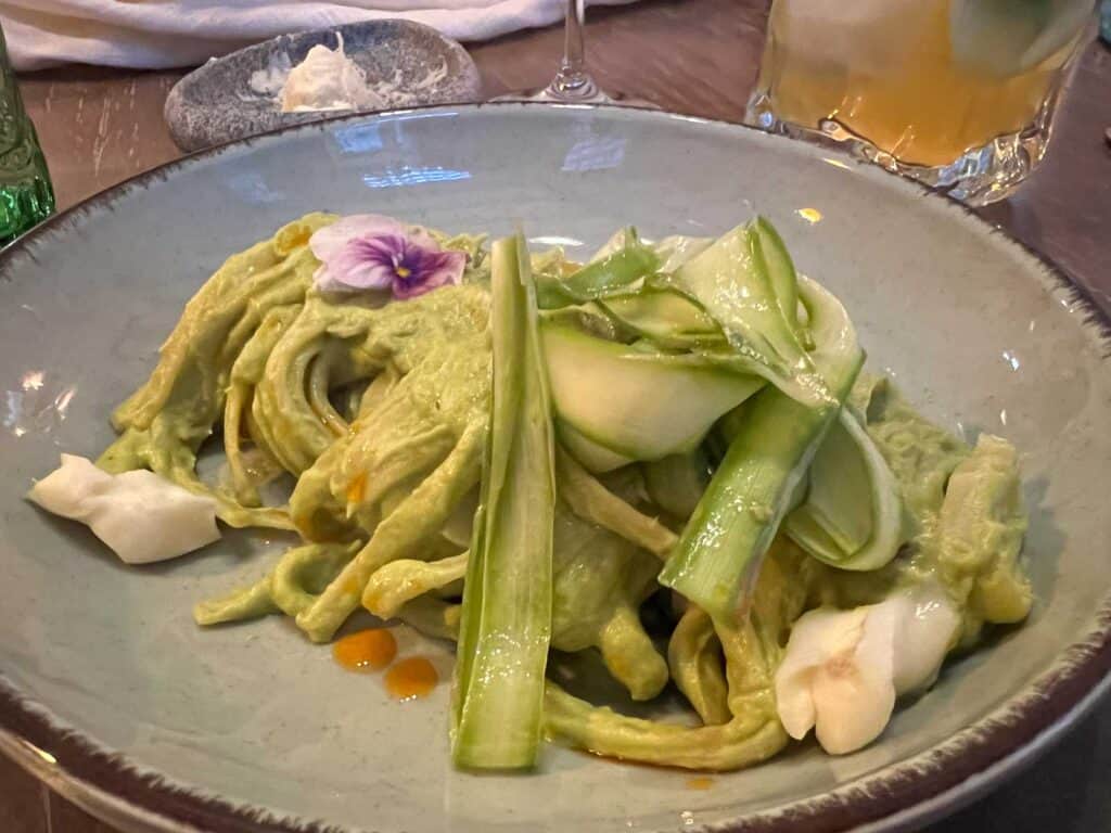 A plate of green pasta topped with zucchini ribbons, garnished with a light-colored flower, and white dollops, served with a side dish and a drink.
