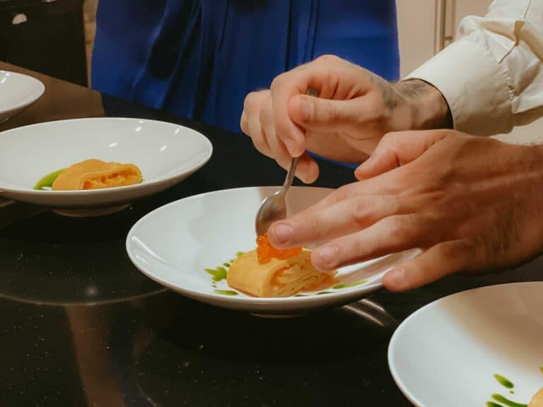 A person garnishes a plated dish with a spoonful of topping in a kitchen setting, with two additional plates of the same dish on the counter.