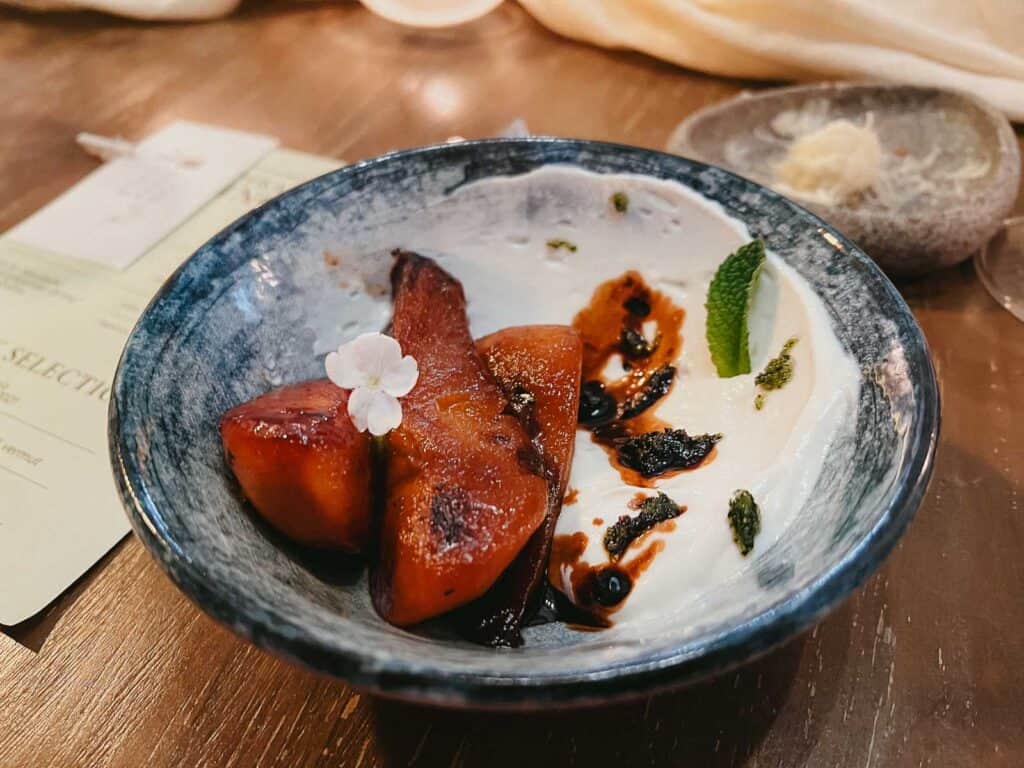 A ceramic bowl with roasted pear, yogurt, mint, balsamic glaze, and garnished with flowers sits on a wooden table with a paper note. Enjoy this local delicacy in the heart of Barcelona through an Eatwith experience.