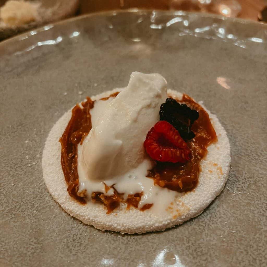 A gourmet dessert featuring a meringue base topped with a scoop of ice cream, caramel sauce, a raspberry, and a garnish, served on a grey plate.