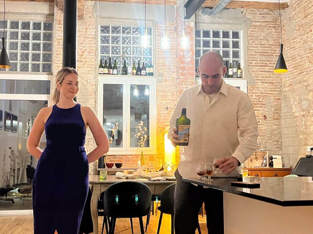 A man pours a drink into a glass at a countertop while a woman in a blue dress stands nearby in a well-lit bar or kitchen with brick walls and large windows.