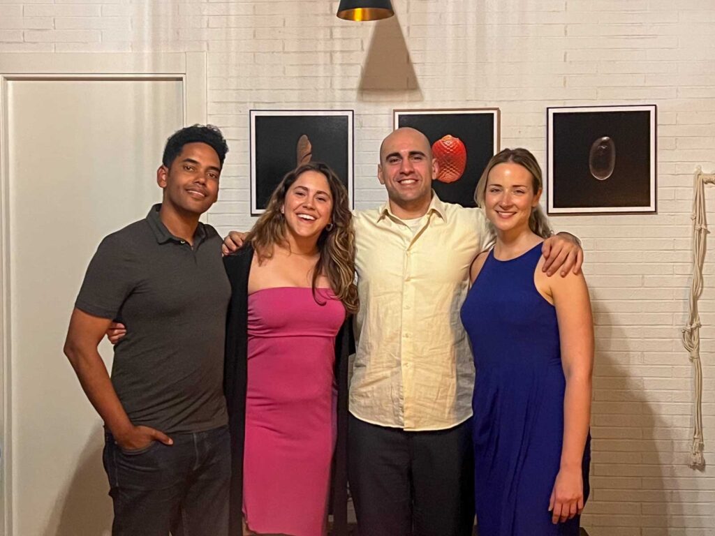 Four people standing side by side with their arms around each other, smiling. They are indoors in front of a white brick wall with framed pictures.
