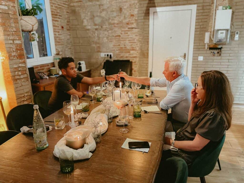 Three people are seated around a table with drinks, candles, and various items. Two are exchanging a bottle while the third person looks on. The rustic setting, complete with exposed brick walls, creates a cozy ambiance ideal for an Eatwith experience in Barcelona.