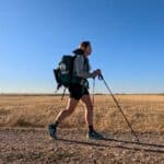 Person hiking with a backpack and trekking poles on a dirt path through a flat, open field under a clear blue sky, reflecting on how they started freelancing and enjoying the freedom that comes with learning how to freelance.