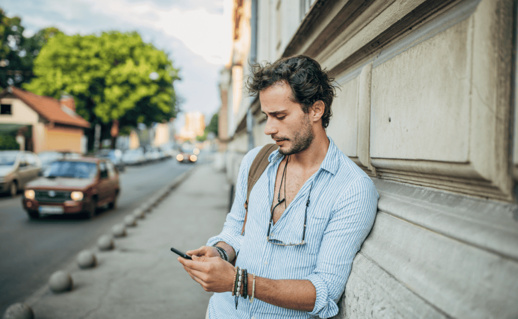 A man with a beard, wearing a striped shirt and bracelets, stands by a building on a city street, intently looking at his phone to read a GigSky review.