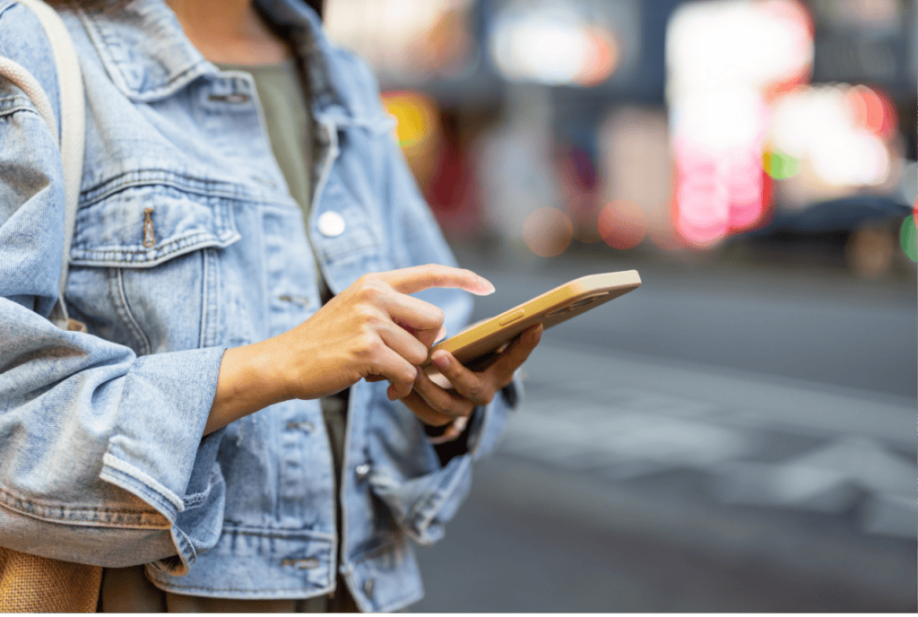 Person wearing a denim jacket, using a smartphone while standing on a city street, possibly reading through a GigSky review.