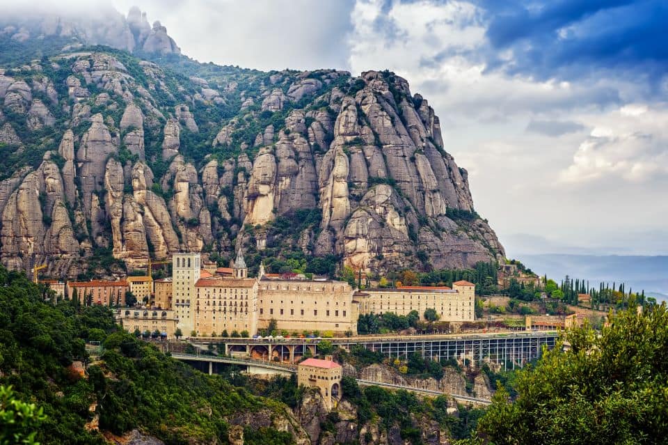 A large monastery is situated at the base of jagged, rocky cliffs with greenery and a cloudy sky overhead, seamlessly blending into your Montserrat itinerary. A bridge is visible in the foreground leading to the entrance.