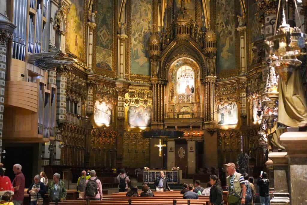 A group of people admire the ornate interior of a richly decorated church featuring a central illuminated shrine, intricate gold details, and seated pews in the foreground—a must-see on any Montserrat itinerary.