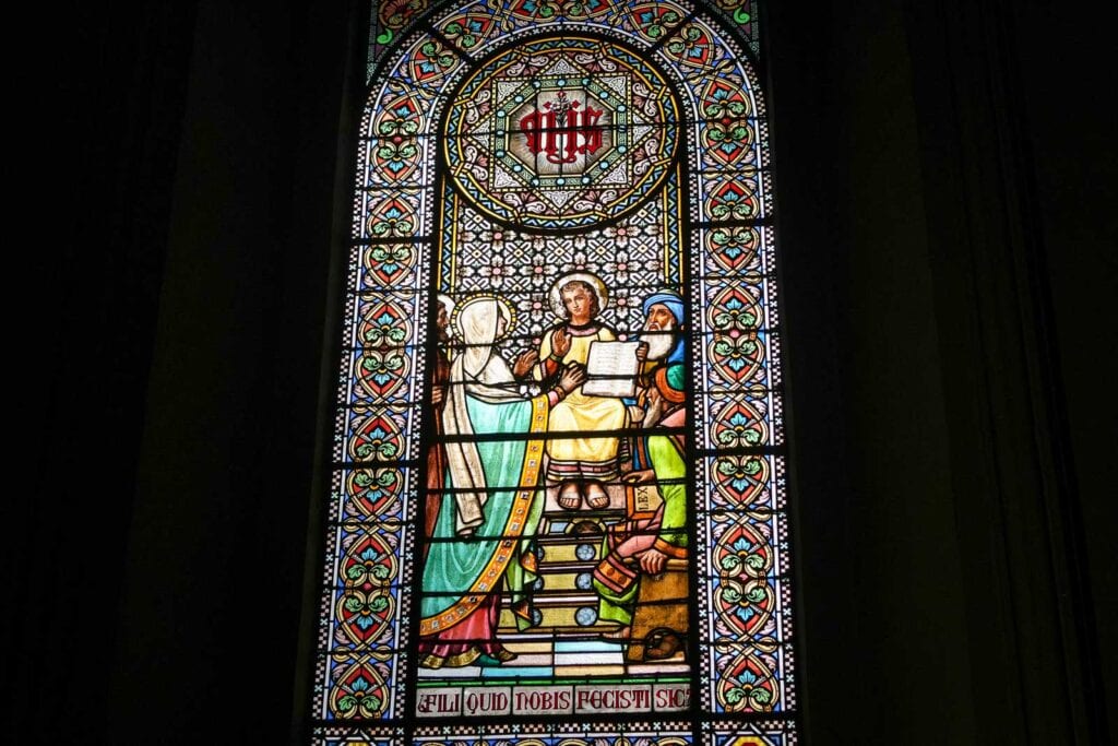 A stained glass window depicts three figures in detailed, vibrant colors — a scene reminiscent of a stop on your Montserrat itinerary. The central figure is seated, holding a book, with two individuals standing on either side. Intricate patterns adorn the window.