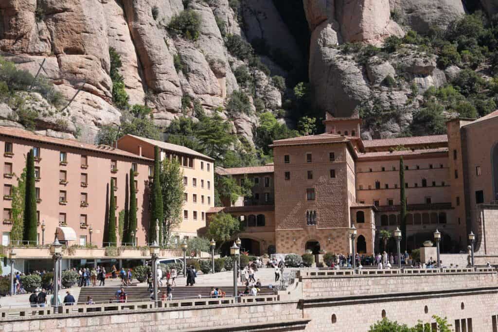 A group of buildings set against a rocky, cliff-like background with tourists strolling around the plaza area in Montserrat, Spain, is a must-see part of any Montserrat itinerary.